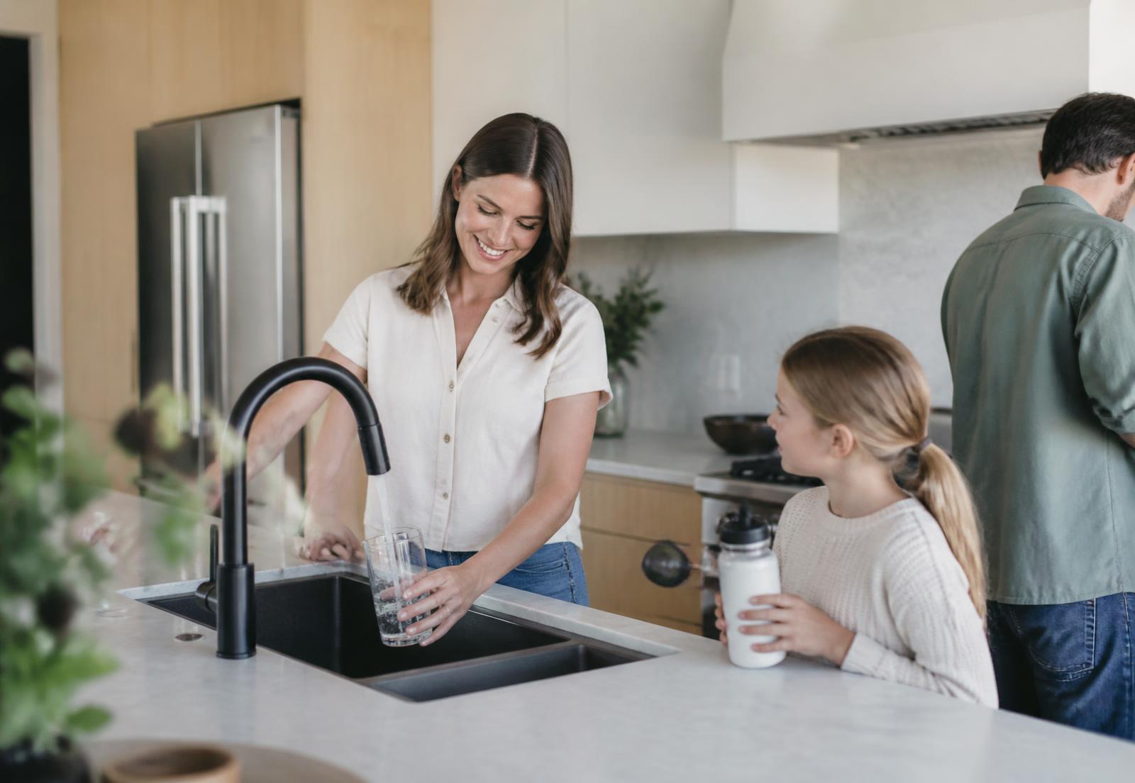 A parent filling a glass of water at a modern kitchen faucet while a child holds a reusable bottle